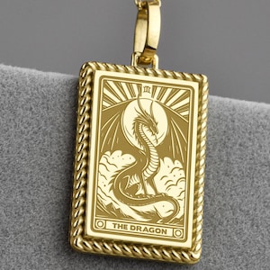 Puede incluir: Collar con colgante dorado con un colgante rectangular con una ilustración detallada de un dragón. El colgante tiene un borde en forma de cuerda y las palabras "THE DRAGON". El collar tiene una cadena delicada con pequeños detalles de bolas.