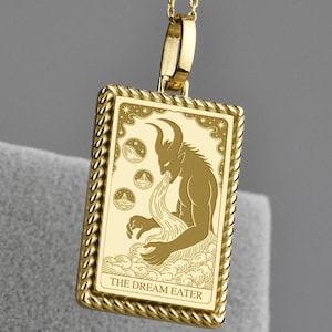Könnte beinhalten: Goldfarbene Anhänger-Halskette mit einem rechteckigen Anhänger mit einer detaillierten Illustration einer gehörnten Figur. Der Anhänger ist mit "THE DREAM EATER" beschriftet und hängt an einer filigranen Kette.