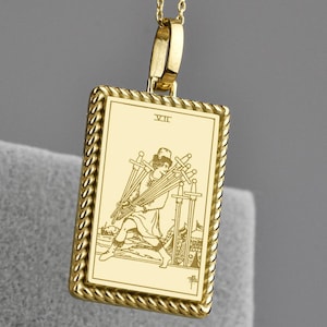 Könnte beinhalten: Goldfarbene Anhängerkette mit einem rechteckigen Anhänger mit einer Tarotkartenillustration. Der Anhänger hat einen seilartigen Rand und die römische Zahl XII. Die Halskette hat kleine, runde Perlen.
