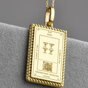 Könnte beinhalten: Eine goldfarbene Halskette mit Anhänger, die einen rechteckigen Anhänger mit detailliertem Design und strukturiertem Rand aufweist. Der Anhänger zeigt Text und Symbole. Die Halskette hat kleine, runde Perlen. Der Anhänger befindet sich vor einem grauen Hintergrund.
