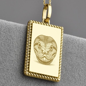 Puede incluir: Collar con colgante dorado con un colgante rectangular con una ilustración detallada de una cabeza de serpiente. El colgante tiene un borde en forma de cuerda y cuelga de una delicada cadena con pequeños detalles de bolas. Sobre fondo gris.