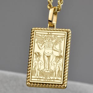 May include: Gold-toned pendant necklace featuring a rectangular pendant with a detailed engraving of a figure with wings and two smaller figures. The pendant is suspended from a rope chain. The text "LE DIABLE" is at the bottom of the pendant.