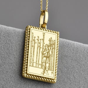Könnte beinhalten: Goldfarbene Halskette mit einem rechteckigen Anhänger, der ein detailliertes graviertes Design aufweist. Der Anhänger zeigt eine Figur, die mehrere Stäbe hält. Der Anhänger hängt an einer filigranen Kette mit kleinen, runden Perlen.