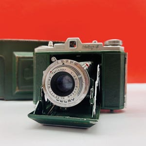 Może przedstawiać: Zabytkowy zielono-srebrny aparat Welmy 6 z skórzanym etui. Aparat posiada obiektyw Terionar z f=75mm i przysłoną 1:4.5. Obiektyw jest otwarty, odsłaniając wewnętrzne elementy. Aparat stoi na czerwonym tle.