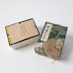 May include: An open box with a beige interior and a lid containing a stack of cards with Japanese text. Beside it, a wrapped package with green and brown patterned paper and Japanese text. The package has a clear plastic wrap.