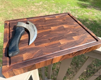 Puede incluir: Una tabla de cortar rectangular de madera con un acabado marrón oscuro y una ranura tallada alrededor del borde. Un cuchillo de carnicero curvo con un mango negro descansa sobre la tabla. La tabla es de madera de testa.