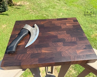 Puede incluir: Una tabla de cortar cuadrada de madera de grano final con un patrón de bloques de color marrón oscuro y tonos variados. Un hacha de metal con una hoja curva y un mango negro descansa sobre la superficie. Se ve un pequeño icono de línea de un gato sosteniendo un cuchillo.