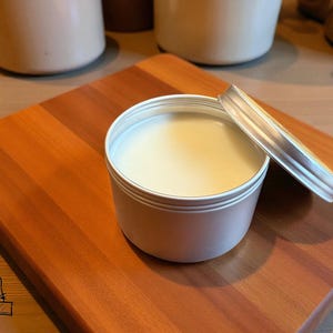 May include: An open, silver-colored tin filled with a creamy, off-white substance, possibly a balm or cream. The tin sits on a wooden cutting board with a reddish-brown hue. Other containers are visible in the background.