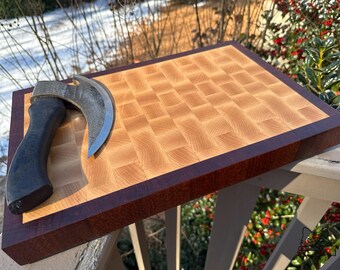 May include: A rectangular butcher block cutting board with a dark brown border and a light wood checkered pattern. A curved blade knife with a black handle rests on the board, suggesting its use for food preparation.