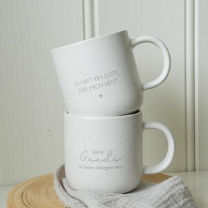 May include: Two white speckled ceramic mugs stacked on a wooden coaster. The top mug has the text "DU BIST EIN GOTT, DER MICH SIEHT." The bottom mug has the text "Seine Gnade ist jeden Morgen neu."