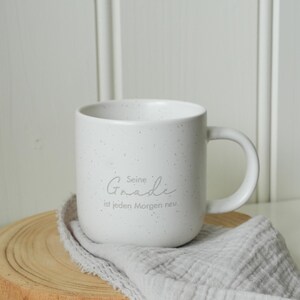 May include: A white speckled ceramic mug with a curved handle. The mug has the text "Seine Gnade ist jeden Morgen neu" in a light gray script. The mug sits on a wooden coaster with a gray cloth draped over it.