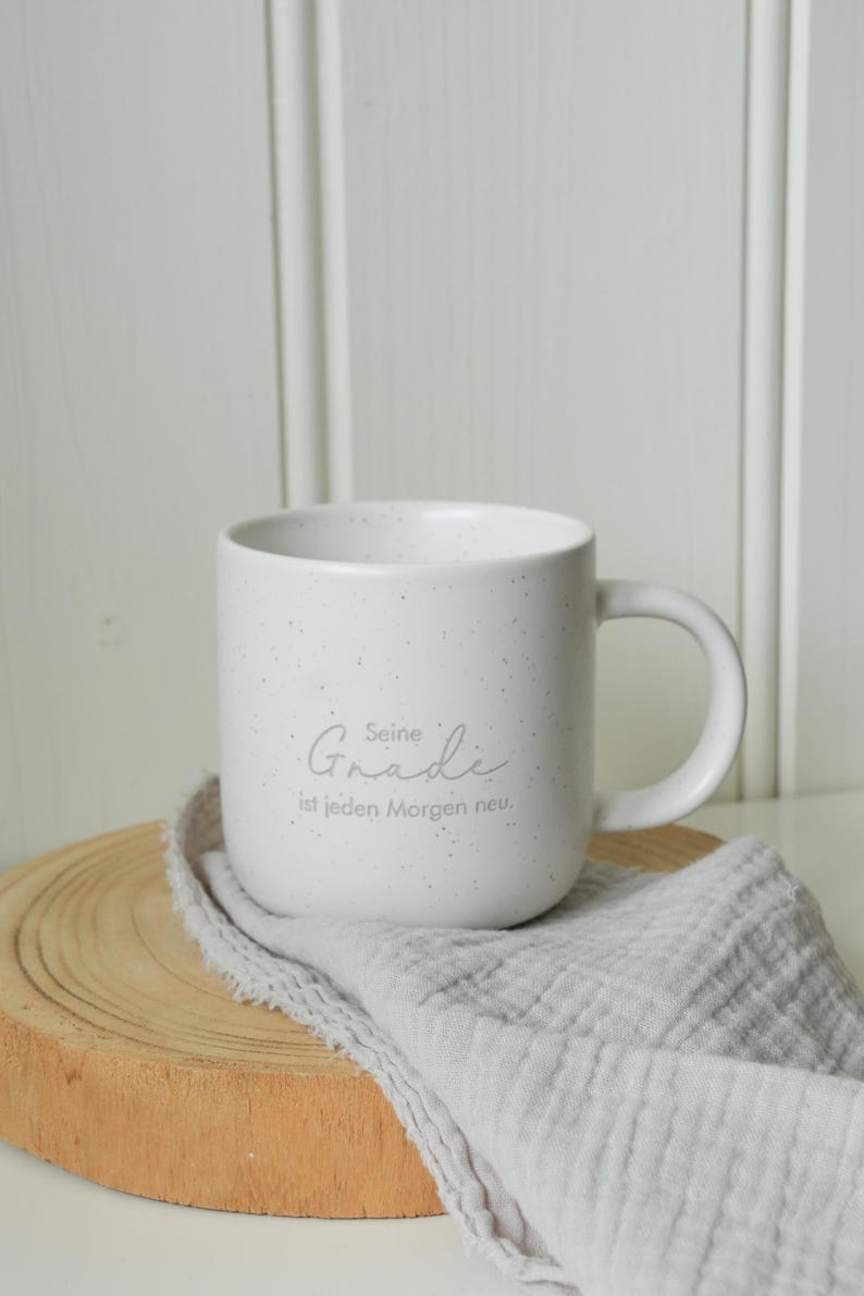 May include: A white speckled ceramic mug with a curved handle. The mug has the text "Seine Gnade ist jeden Morgen neu" in a light gray script. The mug sits on a wooden coaster with a gray cloth draped over it.