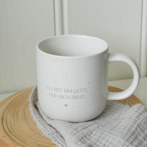 May include: White speckled ceramic mug with a curved handle. The mug has the text "DU BIST EIN GOTT, DER MICH SIEHT." in gray. The mug sits on a wooden slice with a gray cloth.