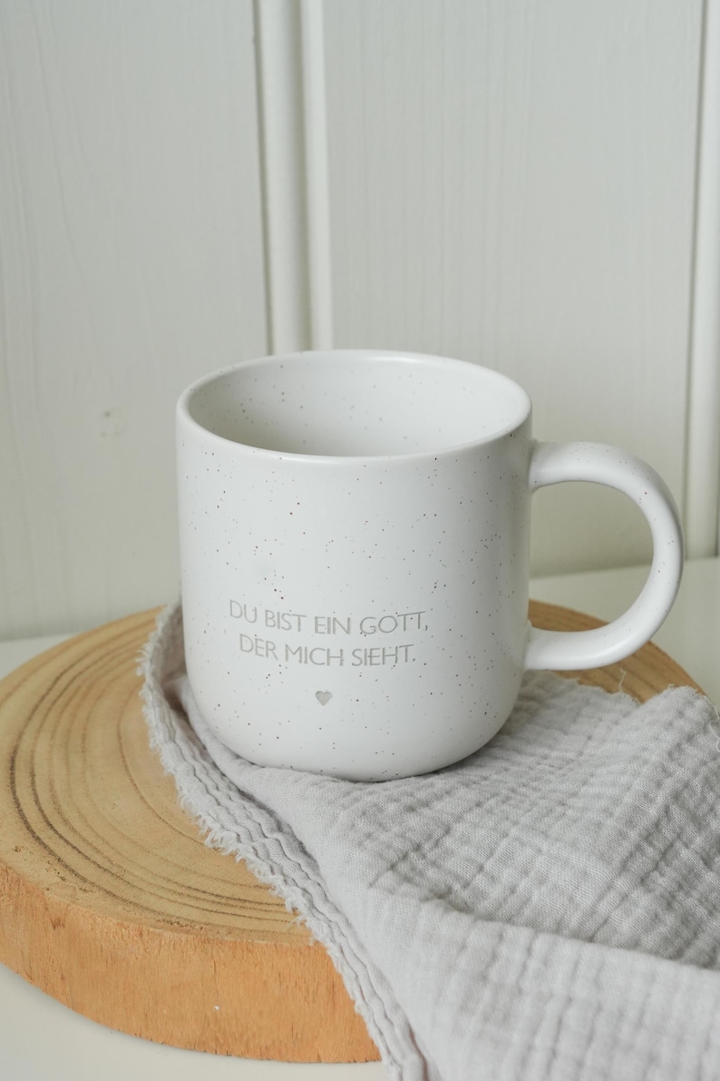 May include: White speckled ceramic mug with a curved handle. The mug has the text "DU BIST EIN GOTT, DER MICH SIEHT." in gray. The mug sits on a wooden slice with a gray cloth.