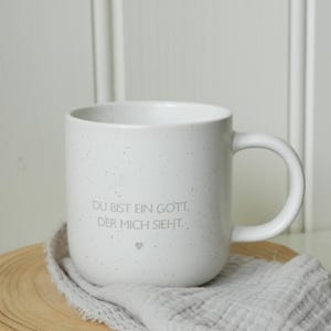 May include: White speckled ceramic mug with a curved handle. The mug has the text "DU BIST EIN GOTT, DER MICH SIEHT." and a small heart. The mug sits on a wooden slice and a gray cloth.
