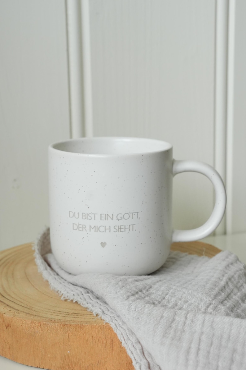 May include: White speckled ceramic mug with a curved handle. The mug has the text "DU BIST EIN GOTT, DER MICH SIEHT." and a small heart. The mug sits on a wooden slice and a gray cloth.