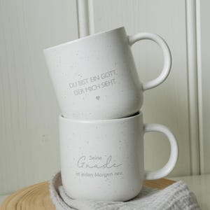 May include: Two white ceramic mugs stacked on top of each other. The top mug has the text "DU BIST EIN GOTT, DER MICH SIEHT." The bottom mug has the text "Seine Gnade ist jeden Morgen neu." The mugs are speckled and have a handle.