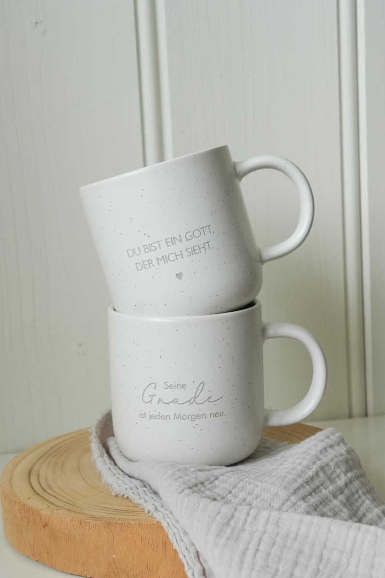 May include: Two white ceramic mugs stacked on top of each other. The top mug has the text "DU BIST EIN GOTT, DER MICH SIEHT." The bottom mug has the text "Seine Gnade ist jeden Morgen neu." The mugs are speckled and have a handle.