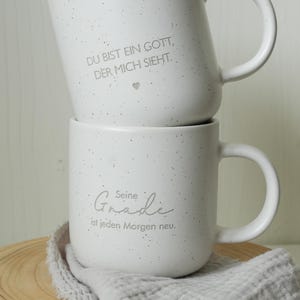 May include: Two white ceramic mugs stacked on top of each other. The top mug has the text "DU BIST EIN GOTT, DER MICH SIEHT." The bottom mug has the text "Seine Gnade ist jeden Morgen neu." Both mugs have a speckled design.