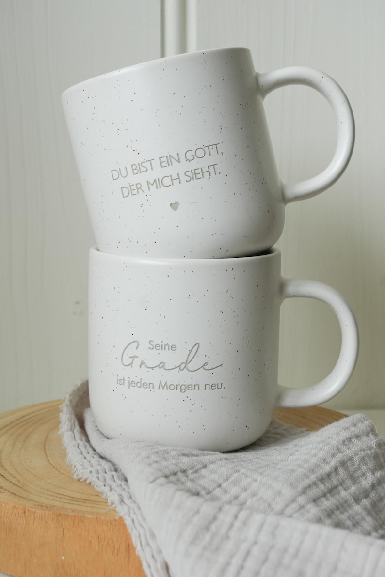 May include: Two white ceramic mugs stacked on top of each other. The top mug has the text "DU BIST EIN GOTT, DER MICH SIEHT." The bottom mug has the text "Seine Gnade ist jeden Morgen neu." Both mugs have a speckled design.
