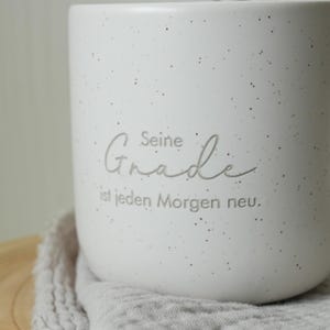 May include: A white speckled ceramic mug with the German text "Seine Gnade ist jeden Morgen neu." The mug has a rounded shape and sits on a gray cloth. The mug is a neutral color and would fit in any kitchen.