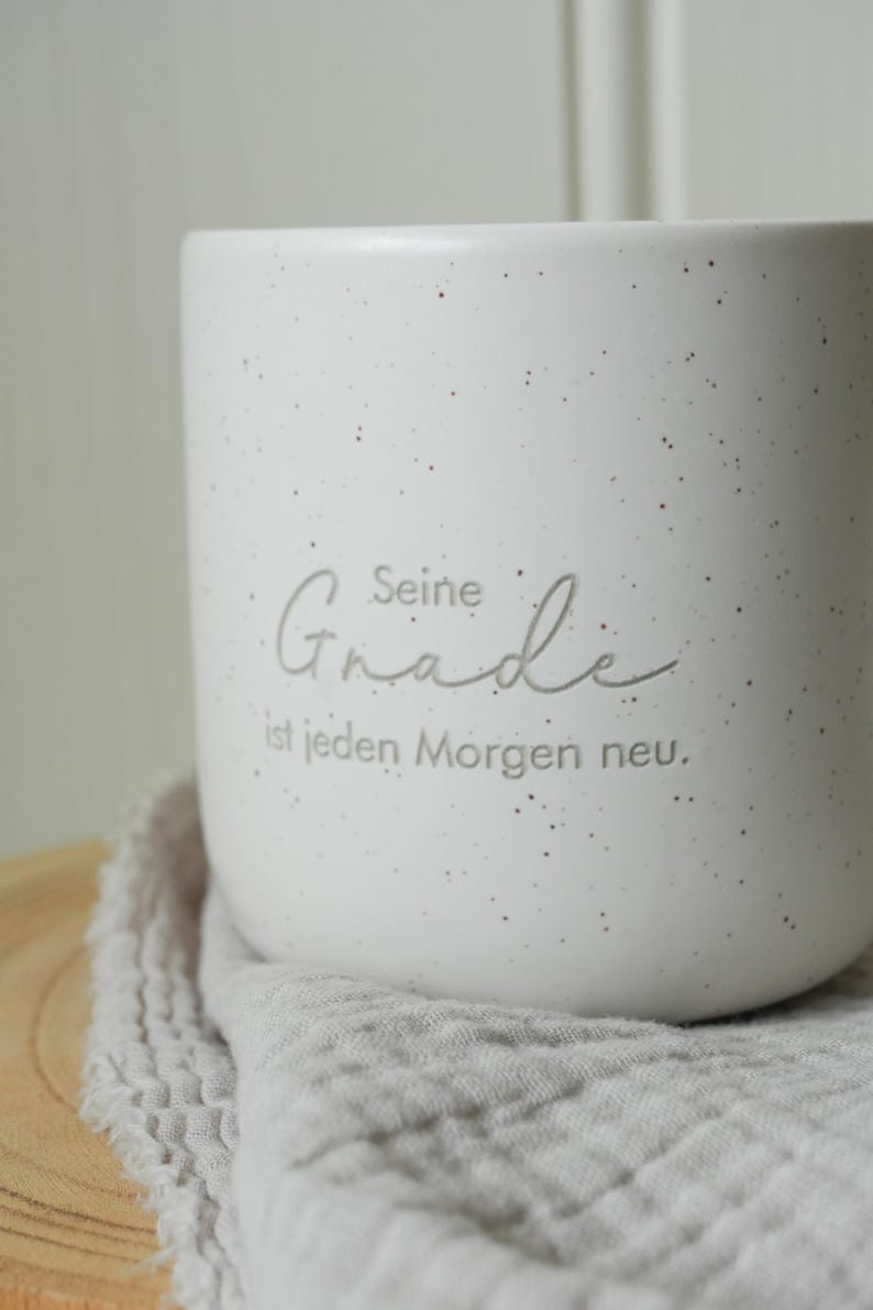 May include: A white speckled ceramic mug with the German text "Seine Gnade ist jeden Morgen neu." The mug has a rounded shape and sits on a gray cloth. The mug is a neutral color and would fit in any kitchen.