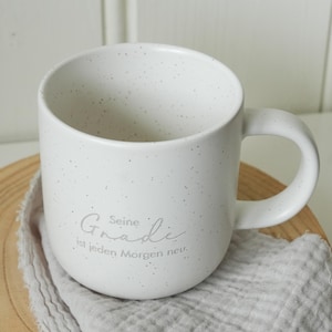 May include: White speckled ceramic mug with a curved handle. The mug has the text "Seine Gnade ist jeden Morgen neu" in a light gray script. The mug sits on a wooden surface with a gray cloth.