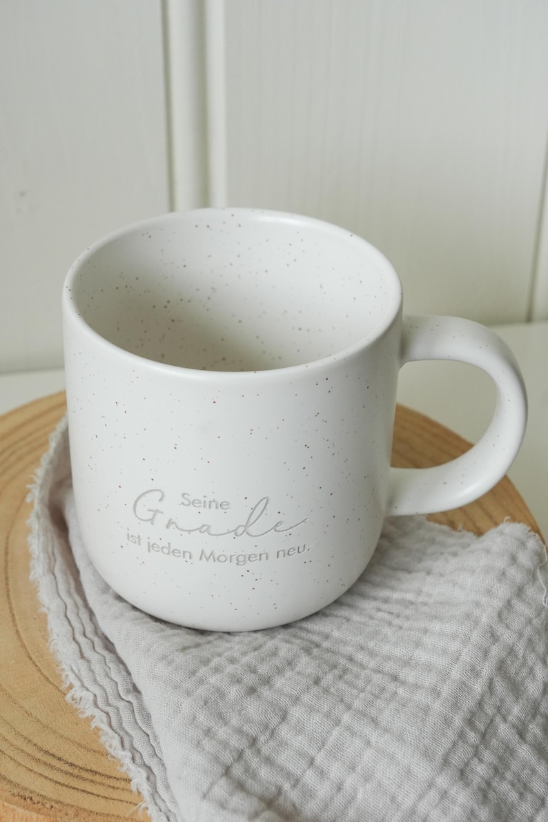 May include: White speckled ceramic mug with a curved handle. The mug has the text "Seine Gnade ist jeden Morgen neu" in a light gray script. The mug sits on a wooden surface with a gray cloth.