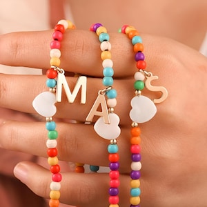 May include: Three colorful beaded bracelets with gold letter charms. The charms spell out "MAS". Each bracelet has white heart-shaped beads.