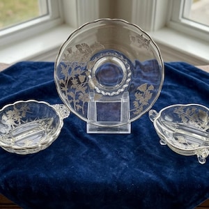 Può includere: Un set da portata in vetro trasparente con disegni floreali argentati. Il set comprende un piatto grande su un supporto trasparente e due piatti più piccoli. I piatti sono su una superficie di velluto blu scuro.