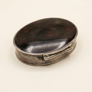 Puede incluir: Una caja antigua de forma ovalada con una tapa oscura y moteada de carey y una base de metal plateado. La caja se cierra con una pequeña bisagra y un cierre. La tapa tiene un acabado brillante y la base tiene un aspecto pulido.