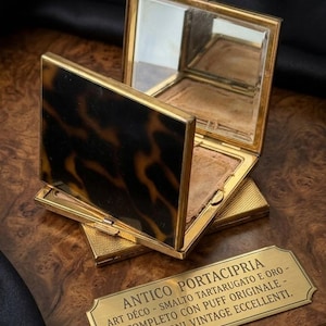 Puede incluir: Un espejo compacto cuadrado de metal dorado con un patrón de carey marrón y negro en la tapa. El compacto está abierto, revelando un espejo interior y una borla beige. Frente a él hay una placa dorada rectangular con texto en italiano.