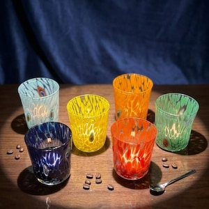 Puede incluir: Un conjunto de seis portavelas de vidrio en varios colores, incluyendo azul, azul claro, amarillo, naranja, rojo y verde. Cada vaso presenta un patrón moteado y jaspeado con pequeños detalles incrustados. Granos de café dispersos y una cuchara de metal.