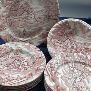 May include: A set of antique white and red transferware plates. The plates depict a detailed rural landscape scene with buildings, trees, and figures. The plates have a scalloped edge and are stacked on a dark blue surface.