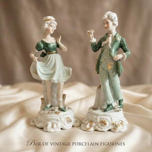 May include: A pair of porcelain figurines depicting two figures in 18th-century attire. One figure wears a green bodice and light skirt, while the other wears a green coat and light trousers. Both stand on ornate white bases with gold accents.