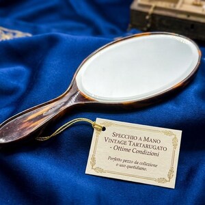 Può includere: Uno specchio a mano ovale con cornice e manico dal motivo tartarugato marrone. Un cartellino rettangolare color crema è attaccato al manico con un cordino dorato, riportante il testo: Specchio a Mano Vintage Tartarugato - Ottime Condizioni.