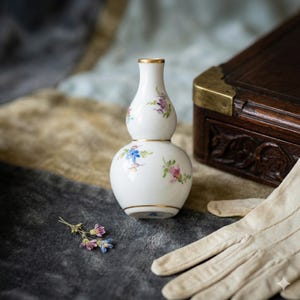 Puede incluir: Un jarrón de porcelana blanco con forma de doble bulbo, adornado con delicados motivos florales en azul, rosa y morado. El jarrón tiene ribetes dorados y está sobre un fondo texturizado, sugiriendo una estética vintage.