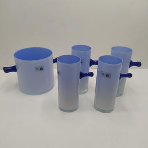 Puede incluir: Un juego de cinco piezas de cristalería azul claro. El juego incluye una jarra grande y redonda y cuatro vasos altos y cilíndricos. Cada pieza tiene un asa azul oscuro y un interior azul claro. La jarra y los vasos tienen una pequeña etiqueta rectangular.