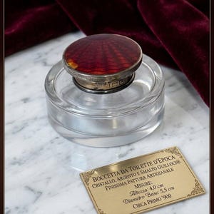 Può includere: Un vasetto da toilette in cristallo trasparente con un coperchio rotondo in argento decorato con smalto guilloché rosso. Una targhetta rettangolare in ottone riporta il testo: BOCCETTA DA TOILETTE D'EPOCA, CRISTALLO, ARGENTO E SMALTO GUILLOCHÉ, FINISSIMA FATTURA ARTIGIANALE, MISURE: Altezza: 4,0 cm, Diametro Base: 5,5 cm, CIRCA PRIMO '900.