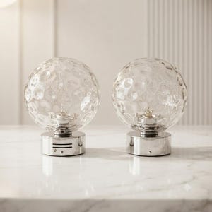 May include: Two spherical table lamps featuring clear, textured glass globes with an irregular, dimpled surface pattern. Each lamp is supported by a cylindrical, polished silver-tone metal base with horizontal ventilation slits near the bottom.