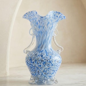 Puede incluir: Un jarrón de cristal transparente con un patrón moteado en azul y blanco por todo el cuerpo. El jarrón tiene un borde superior con volantes, dos asas curvas de cristal transparente a los lados y una base decorativa festoneada.