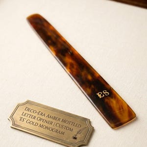 May include: A Deco-Era amber mottled letter opener with a custom 'ES' gold monogram. The opener is a long, slender shape with a tortoiseshell pattern. A brass plaque next to it reads "DECO-ERA AMBER MOTTLED LETTER OPENER | CUSTOM 'ES' GOLD MONOGRAM."