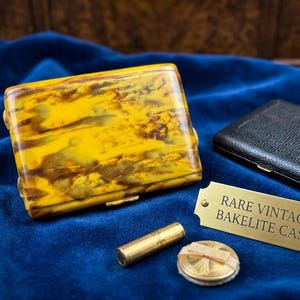 May include: A rectangular yellow and brown marbled case, a black textured case, a small gold-toned cylindrical tube, a round gold-toned compact with a ribbon detail, and a brass plaque with the text RARE VINTAGE BAKELITE CASE.
