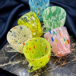 Könnte beinhalten: Ein Set aus sechs Glastassen mit einem gesprenkelten Muster in verschiedenen Farben wie Hellblau, Grün, Gelb, Creme und Rosa. Jede Tasse hat einen kleinen Henkel und eine glatte, glänzende Oberfläche. Der Text Original Murano Art Glass Set of 6 ist sichtbar.