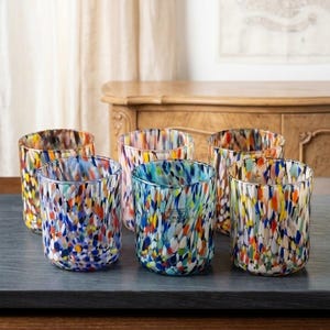 Könnte beinhalten: Ein Set aus sechs zylindrischen Glasbechern mit einem mehrfarbigen Konfetti-Muster aus blauen, orangen, gelben und weißen Sprenkeln. Jedes Gefäß hat einen glatten, abgerundeten Rand und einen klaren Boden, der die lebendige, gesprenkelte Glaskunst zeigt.