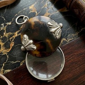 May include: A round antique magnifying glass with a tortoiseshell cover and silver accents. The magnifying glass is open, revealing the clear glass lens. The cover has decorative silver embellishments and a small loop for a chain.
