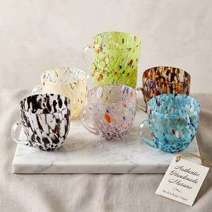 Könnte beinhalten: Ein Set aus sechs Glastassen mit gesprenkelten Mustern in Limettengrün, Creme, Schwarz, Rosa, Braun und Blau. Jede Tasse hat einen Henkel aus klarem Glas. Ein weißes Etikett trägt den Text Authentic Handmade Murano Set di 6 Tazze Unique.