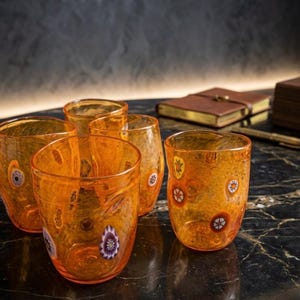 Könnte beinhalten: Ein Set aus vier orangefarbenen Glasbechern mit gesprenkelter Textur und eingebetteten Millefiori-Mustern in verschiedenen Farben wie Violett, Weiß und Gelb. Jeder Becher hat eine abgerundete, leicht unregelmäßige Form und eine glatte Oberfläche.