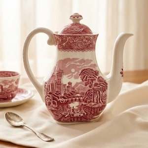 Peut inclure: Une théière en céramique de couleur crème ornée d'un motif détaillé en transfert bordeaux représentant un paysage avec un château, un pont et des personnages dans une barque. La théière possède un couvercle assorti, une anse et un long bec.