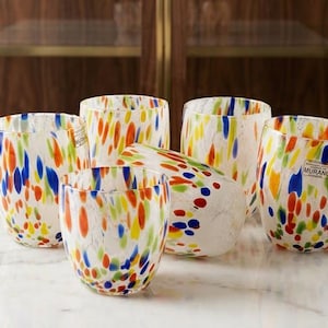 Puede incluir: Un juego de cinco vasos de cristal transparente con un colorido patrón de confeti de motas rojas, azules, naranjas y amarillas. Cada vaso redondeado tiene un acabado liso y una pequeña pegatina en el lateral que dice Murano, indicando su origen.